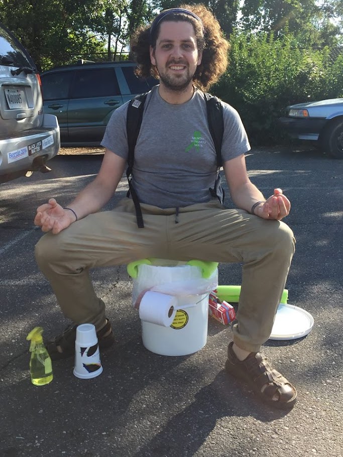 Picture of me in 2018 sitting on a homemade bucket toilet and toileting supplies in church parking lot before passking kit to homeless encampers by river.